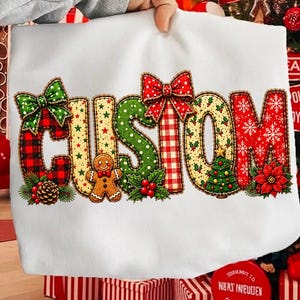 May include: A white sweatshirt featuring the word CUSTOM in large, decorative letters. Each letter has a unique holiday pattern, including red and black plaid, green polka dots, red gingham, and snowflakes, accented with bows, a gingerbread man, and a tree.