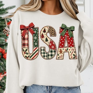 Könnte beinhalten: Ein cremefarbenes Sweatshirt mit Rundhalsausschnitt und dem Schriftzug USA im Patchwork-Design. Die Buchstaben sind mit verschiedenen Weihnachtsmustern verziert, darunter Karos, Schneeflocken, Tannenbäume, Leopardenmuster, Lebkuchenmänner und Zuckerstangen, sowie Schleifen.