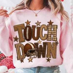 May include: A light pink crewneck sweatshirt featuring the text TOUCH DOWN in large, textured brown letters with various patterns including plaid, herringbone, and cable knit. A brown football and a brown football helmet are integrated into the design.