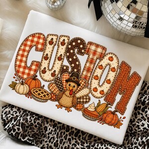 Può includere: Una t-shirt bianca con la parola CUSTOM in grandi lettere decorate. Le lettere presentano motivi a quadri arancioni, pois e zucche. Un tacchino in stile cartoon con un cappello da pellegrino è circondato da zucche, una torta e pannocchie di mais.