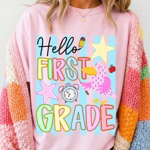 May include: A light pink t-shirt featuring a graphic with the text Hello FIRST GRADE in colorful letters. The design includes a yellow pencil, a yellow star, a pink star, a blue alarm clock, a red apple, and a pink bird holding a pointer stick.