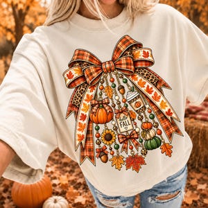 Pode incluir: Uma t-shirt cor de creme com um grande gráfico de um laço com padrão xadrez e leopardo. Do laço pendem correntes com berloques que incluem abóboras, um girassol, bolotas, um copo de café e uma etiqueta com o texto hello FALL.