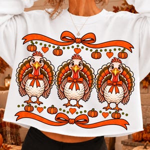 May include: A white long-sleeve sweatshirt featuring a graphic print of three turkeys, each wearing an orange bow tie. The design includes orange ribbons with bows at the top and bottom, interspersed with small orange pumpkins and orange hearts.
