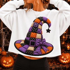 May include: A white long-sleeve sweatshirt featuring a large, central graphic of a patchwork witch hat. The hat is composed of quilted squares in orange, purple, and black, decorated with star and bat patterns, and includes a purple bow.