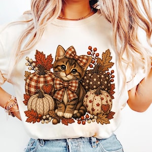 May include: A cream-colored t-shirt featuring a graphic print of a brown tabby kitten wearing a plaid bow on its head and neck. The kitten is surrounded by four pumpkins with various patterns, autumn leaves, and small berries in shades of brown and orange.