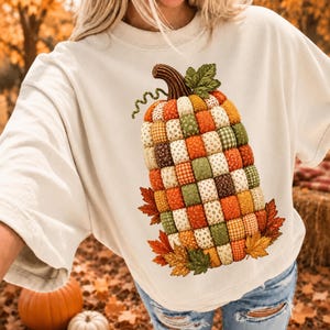 Op de afbeelding: Een crèmekleurig sweatshirt met ronde hals en een grote afbeelding van een pompoen gemaakt van een patchwork van diverse stoffen vierkantjes in tinten oranje, groen en geel, geaccentueerd met een bruine steel, een groen blad en herfstbladeren.