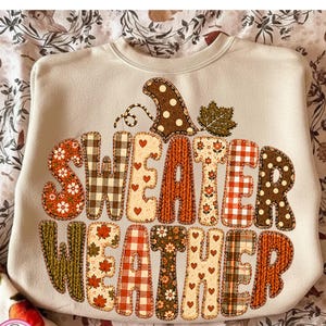 Puede incluir: Una sudadera de cuello redondo en color beige con un gran gr&aacute;fico en forma de calabaza que contiene el texto "SWEATER WEATHER". Las letras est&aacute;n decoradas con varios patrones que incluyen cuadros, flores, lunares y texturas de punto en tonos oto&ntilde;ales.