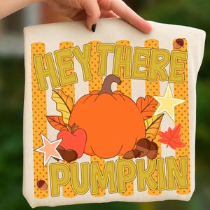 Op de afbeelding: Een crèmekleurige tote bag met een grafische print met de tekst HEY THERE PUMPKIN in het geel. Het ontwerp bevat een grote oranje pompoen, een rode appel, bruine eikels, gele en rode herfstbladeren en sterren op een gele gestreepte achtergrond.