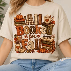 May include: A beige short-sleeve t-shirt featuring a graphic print with the text All Booked for Fall. The design includes stacks of books, pumpkins, a mug with a heart, eyeglasses, and autumn leaves in shades of orange, brown, and cream.