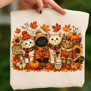 May include: A cream-colored t-shirt featuring a graphic of four kittens wearing autumn-themed clothing, including knit hats and scarves. The design includes orange pumpkins, sunflowers, maple leaves, and a mug labeled Pumpkin Spice with Hello Fall text.