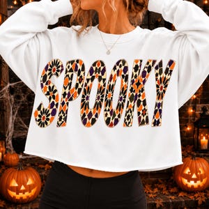 May include: A white long-sleeve cropped sweatshirt featuring the word SPOOKY printed across the chest. The letters are filled with a geometric pattern in shades of orange, purple, black, and gold, creating a vibrant and textured visual effect.