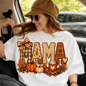 May include: A white crewneck sweatshirt featuring the word MAMA in large, textured letters with various autumn-themed patterns including plaid, cable knit, and fall leaves. The design includes a plaid bow, pumpkins, and a mug of hot cocoa.