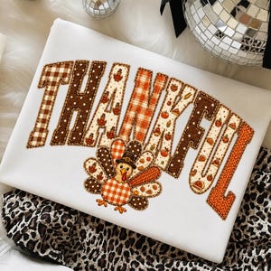May include: A white sweatshirt featuring the word THANKFUL in large, block-style letters. Each letter has a unique pattern including brown and white gingham, polka dots, orange plaid, and knit textures, accented with small turkey and pumpkin icons.
