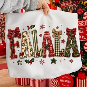 Può includere: Una felpa bianca con la scritta FALALA in grandi lettere decorative. Ogni lettera presenta un motivo natalizio unico, tra cui tartan rosso, fiori color crema, strisce verdi, maglia rossa e agrifoglio, decorata con fiocchi e stelle.