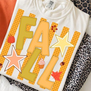 以下が含まれることがあります： クリーム色のTシャツで、黄色とオレンジ色の大きなブロック体で「FALL」という文字がプリントされています。デザインには、オレンジ色の水玉模様の縦縞、小さなオレンジ色のカボチャ、カップケーキ、赤い葉、どんぐり、2つの星が含まれています。