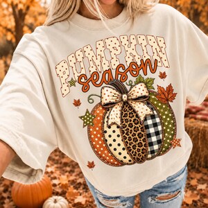 Puede incluir: Una camiseta color crema con un gráfico de calabaza con segmentos de lunares naranjas, estampado de leopardo y cuadros vichy en blanco y negro. El texto PUMPKIN season aparece sobre la calabaza, decorado con pequeñas hojas de arce y estrellas.