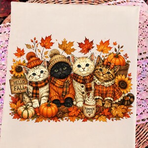 May include: A white fabric towel featuring four kittens wearing autumn clothing, surrounded by orange maple leaves, sunflowers, and pumpkins. A wooden sign reads HELLO FALL and a mug is labeled PUMPKIN SPICE with a small heart icon below the text.