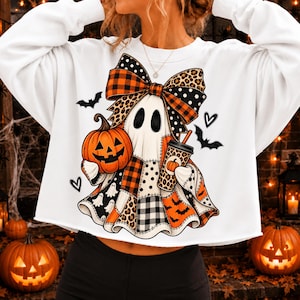 Puede incluir: Una sudadera blanca de manga larga con el gráfico de un fantasma que lleva un lazo grande. El fantasma sostiene una calabaza naranja y un vaso de café. El diseño incluye patrones de cuadros, estampado de leopardo, lunares y murciélagos.