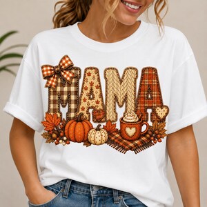 May include: A white t-shirt featuring the word MAMA in large, decorative letters with various autumn-themed patterns including plaid, cable knit, and leaf prints. The design includes a plaid bow, orange and white pumpkins, and a mug of cocoa.