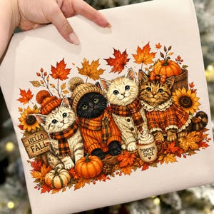 May include: A light-colored fabric featuring a colorful illustration of four kittens wearing autumn clothing, surrounded by orange maple leaves, sunflowers, and pumpkins. A wooden sign reads HELLO FALL and a mug is labeled PUMPKIN SPICE.