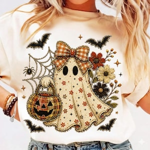 May include: A cream-colored t-shirt featuring a graphic of a ghost wearing a brown and white checkered bow. The ghost holds a checkered pumpkin bucket near a spider web with a spider, surrounded by black bats and colorful autumn flowers.