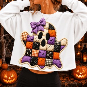May include: A white long-sleeve sweatshirt featuring a large, ghost-shaped applique made of quilted fabric squares in orange, purple, black, and white patterns. The ghost has a purple floral bow and is outlined with small wooden beads.