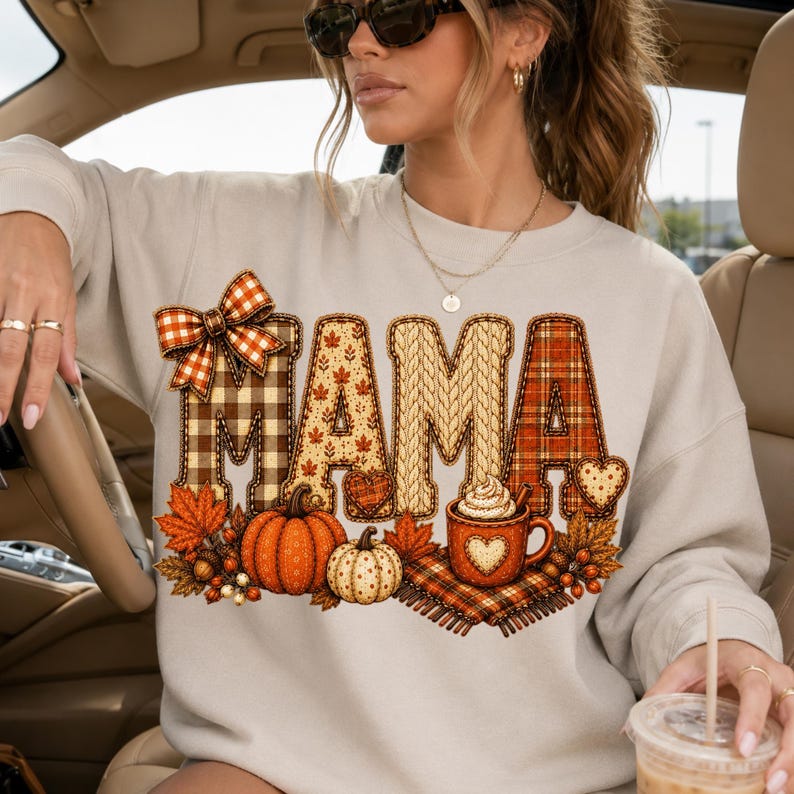 May include: A beige long-sleeve sweatshirt featuring the word MAMA in large, patterned letters. The design includes a plaid bow, autumn leaves, pumpkins, and a mug of hot cocoa with whipped cream, all in shades of orange, brown, and cream.