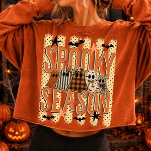 Könnte beinhalten: Ein langärmeliges Sweatshirt in gebranntem Orange mit einem Grafikdruck, der den Text "SPOOKY SEASON" in großen orangefarbenen Buchstaben zeigt. Das Design umfasst fünf stilisierte Geister mit verschiedenen Mustern, kleine schwarze Fledermäuse und Sterne.