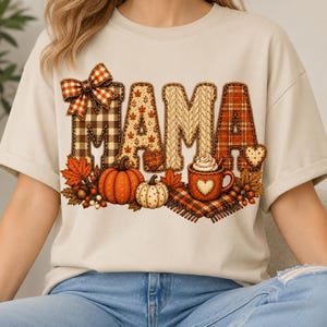 May include: A beige short-sleeve t-shirt featuring the word MAMA in large letters with varied autumn-themed patterns including plaid, cable knit, and leaves. The design includes a plaid bow, pumpkins, a mug of cocoa, and fall foliage.