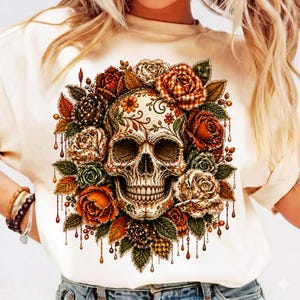 Peut inclure: Un t-shirt couleur crème arborant un motif central représentant un crâne humain entouré d'un arrangement dense de roses et de feuilles dans des tons orange, rouge, marron et vert. Le crâne présente des lignes et motifs décoratifs.