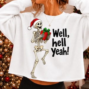 Puede incluir: Una sudadera blanca de manga larga y corte corto con el gráfico de un esqueleto que lleva un gorro de Papá Noel rojo y sostiene un regalo rojo con un lazo verde. El texto "Well, hell yeah!" está impreso en negro junto al esqueleto.