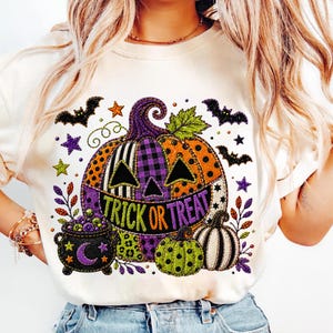 May include: A cream-colored t-shirt featuring a large graphic of a multi-patterned pumpkin with the text TRICK OR TREAT. The design includes a black cauldron with purple bubbles, a crescent moon, small bats, stars, and a smaller green pumpkin.