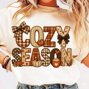 May include: A cream-colored t-shirt featuring the text COZY SEASON in large, decorative letters. The letters are filled with various patterns including brown plaid, cable knit, polka dots, and autumn leaves, accented by bows, a pumpkin, and a mug.