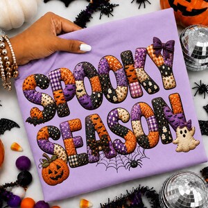 Może przedstawiać: Jasnofioletowy t-shirt z napisem SPOOKY SEASON wykonanym czcionką w stylu patchworkowym. Litery wypełnione są pomarańczowymi, fioletowymi i czarnymi wzorami, w tym duchami, nietoperzami i kratką. Nadrukowano również małą dynię i duszka.