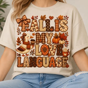 May include: A beige t-shirt featuring a graphic with the text FALL IS MY LOVE LANGUAGE. The design includes a duck wearing a scarf and bow, a pumpkin, a coffee mug, and autumn leaves, all rendered in a patchwork pattern of orange and brown.