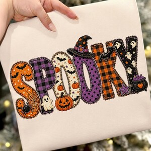 May include: A cream-colored fabric featuring the word SPOOKY in large, appliqued letters. Each letter has a unique pattern including orange spiderwebs, purple plaid, pumpkins, ghosts, bats, a witch hat, orange plaid, and a black cauldron.
