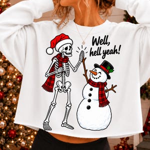 May include: A white long-sleeve sweatshirt featuring a graphic of a skeleton wearing a red Santa hat and a red plaid scarf high-fiving a snowman with a black top hat and a red plaid scarf. The text "Well, hell yeah!" is printed in black.