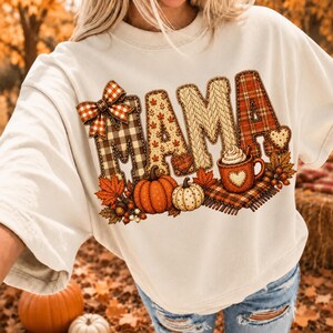 May include: A cream-colored crewneck sweatshirt featuring a large graphic print of the word MAMA in varied autumn-themed patterns including plaid, knit, and leaf prints. The design includes a plaid bow, pumpkins, and a mug of hot cocoa.