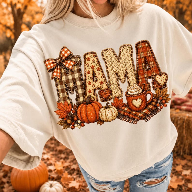 May include: A cream-colored crewneck sweatshirt featuring a large graphic print of the word MAMA in varied autumn-themed patterns including plaid, knit, and leaf prints. The design includes a plaid bow, pumpkins, and a mug of hot cocoa.