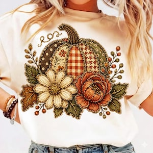 May include: A cream-colored t-shirt featuring a central graphic of a pumpkin composed of various patterned segments including plaid, polka dots, and houndstooth, surrounded by orange and white flowers, green leaves, and small berries.