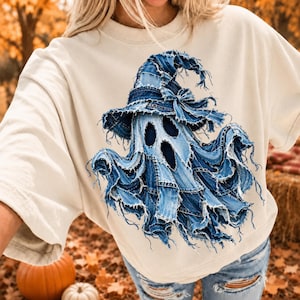 May include: A cream-colored short-sleeve t-shirt featuring a large graphic of a ghost wearing a witch hat. The ghost and hat are composed of a blue denim patchwork design with frayed, raw edges, creating a textured, layered appearance.