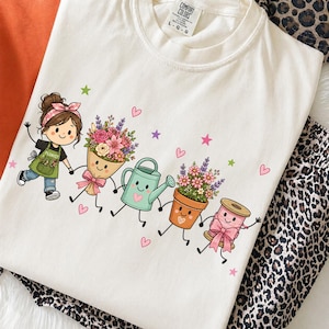 May include: A cream-colored t-shirt featuring a graphic of a cartoon florist, a bouquet of flowers, a light green watering can, a potted plant, and a pink spool of thread, all holding hands. The design includes small pink hearts and stars.