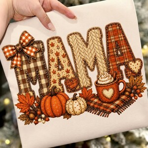 May include: A white fabric surface featuring a graphic print of the word MAMA in large letters with varied textures including plaid, autumn leaves, and cable knit patterns. A plaid bow, orange and white pumpkins, and a mug of cocoa are included.