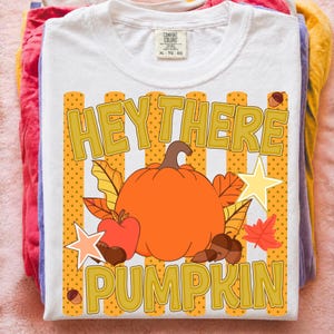May include: A white short-sleeve t-shirt featuring a graphic with the text "HEY THERE PUMPKIN" in gold letters. The design includes a large orange pumpkin, a red apple, brown acorns, autumn leaves, and two light yellow stars on a dotted background.