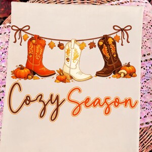 May include: A light-colored t-shirt featuring a graphic of three cowboy boots in orange, white, and brown hanging from a string decorated with autumn leaves and acorns. Orange pumpkins sit below the boots, and the text Cozy Season is printed below.