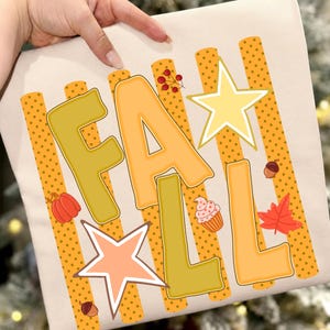 May include: A beige t-shirt featuring the word FALL in large, blocky, yellow and orange letters. The design includes vertical orange polka-dot stripes, a yellow star, a peach star, a red pumpkin, a cupcake, a red leaf, and small acorns.