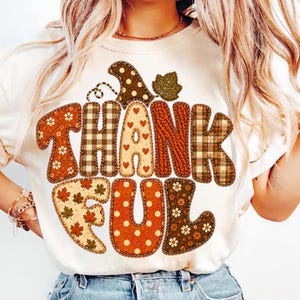 May include: A cream-colored t-shirt featuring the word THANKFUL in large, blocky letters. Each letter has a unique pattern, including plaid, floral, polka dots, hearts, and cable knit textures, topped with a brown polka-dot pumpkin stem and leaf.