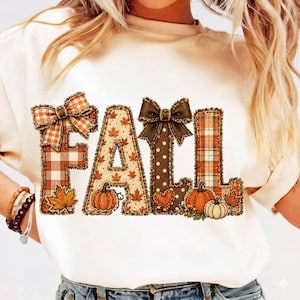 Puede incluir: Una camiseta de color crema con la palabra FALL en letras grandes. Cada letra tiene un patrón único que incluye cuadros naranjas, hojas de otoño, lunares marrones y rayas naranjas, decorada con lazos marrones y naranjas.