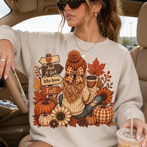 May include: A beige crewneck sweatshirt featuring a graphic print of a person with long hair wearing a knit hat and bow, sitting among pumpkins, sunflowers, and autumn leaves. A wooden signpost reads Just A Girl Who Loves Fall in brown text.