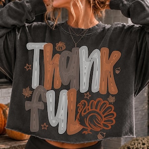 May include: A charcoal grey long-sleeve sweatshirt with a raw hem featuring the text Thankful in a white and orange line-art font. The design includes graphic illustrations of a turkey, a pumpkin, acorns, maple leaves, and small stars.